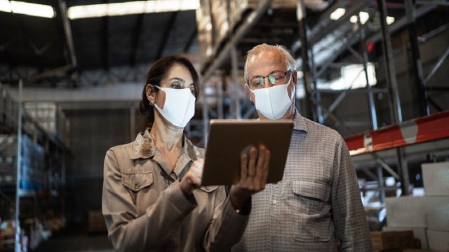 Senior partners walking and using digital tablet at warehouse - with face mask